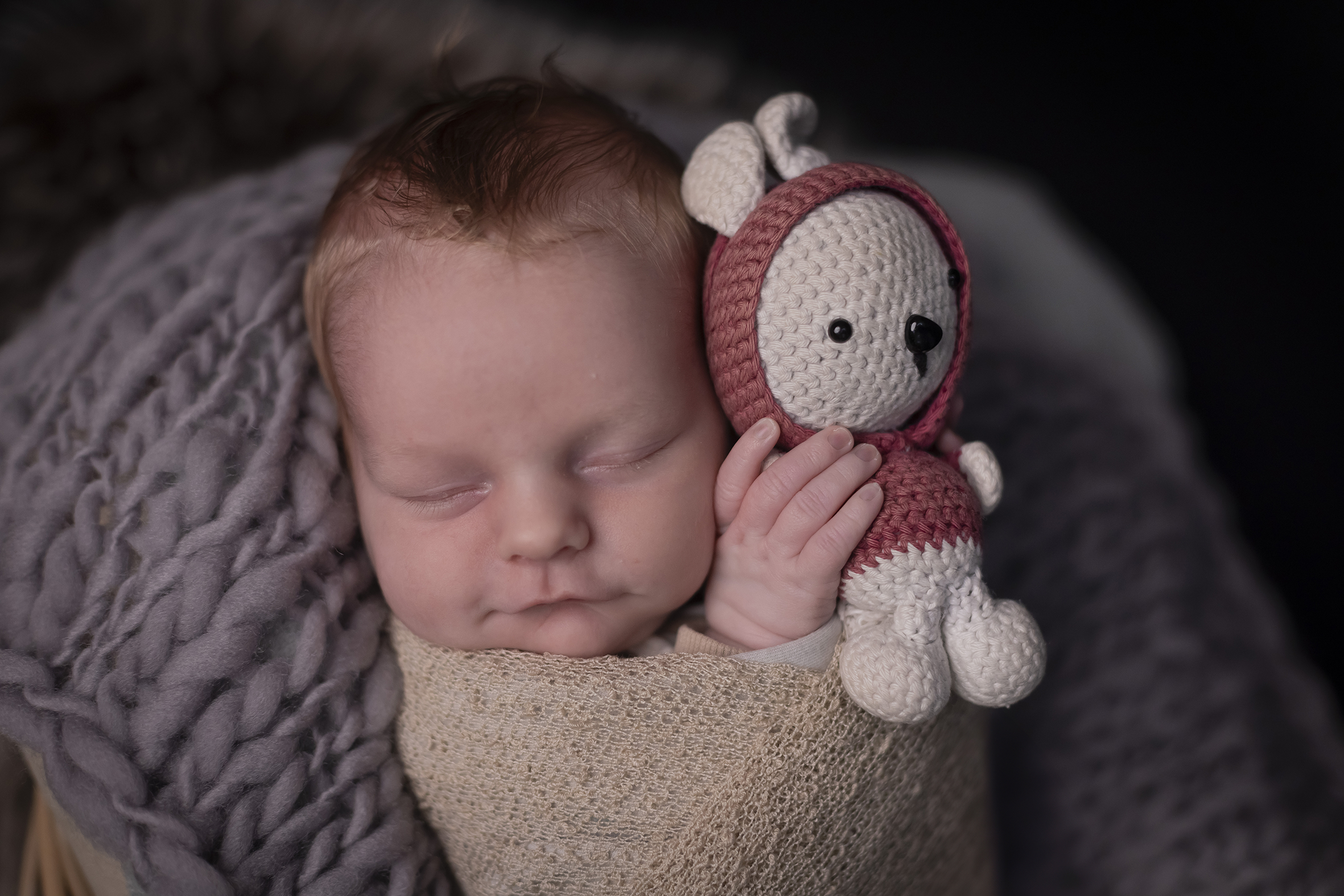 Newborn Baby liegt eingepackt in einer sandfarbenen Decke in einem Korb. Der Korb ist mit einer grobmaschigen grauen Decke ausgelegt,. Das Baby lächelt sanft und hat in seinen Händen einen kleinen Stofftierhasen