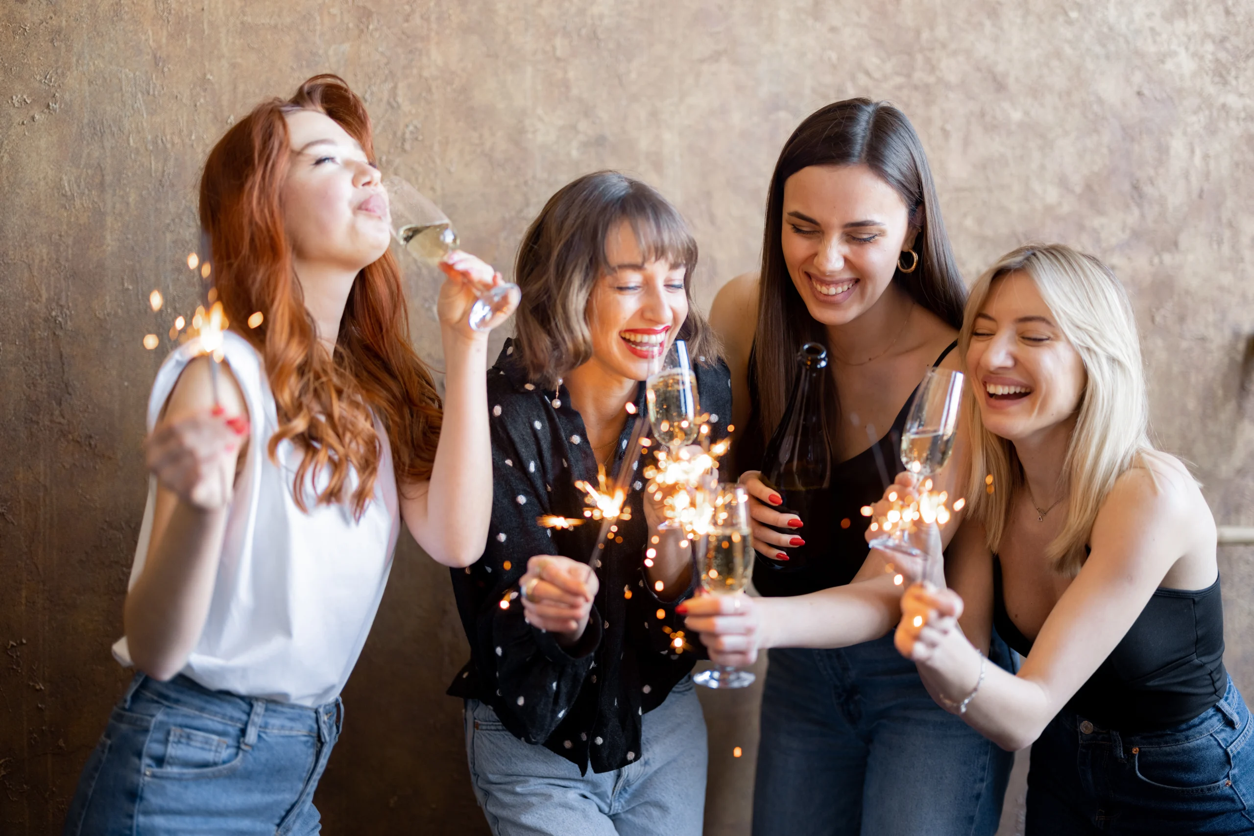 Vier junge Frauen feiern gemeinsam mit Sektgläsern und Wunderkerzen in der Hand. Sie lachen ausgelassen, stoßen an und genießen den Moment. Eine von ihnen hält zudem eine geöffnete Flasche. Der Hintergrund ist eine stilvolle, strukturierte Wand.